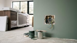 Exposed electrical wires sticking out of an unfinished hole in a freshly painted sage green wall next to paint cans and brushes.