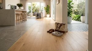 Open-concept home hallway featuring natural light oak timber flooring transitioning with a slim brass threshold trim to grey porcelain kitchen tiles on the left and geometric outdoor patio tiles on the right.