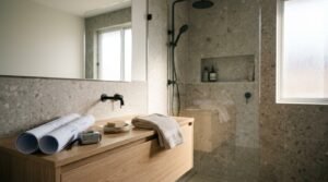 Modern Scandinavian ensuite bathroom featuring a floating oak timber vanity with rolled blueprints, tape measure, wall-mounted matte black tapware, grey terrazzo wall and floor tiles, and a walk-in shower with an integrated niche.