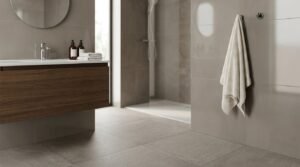Modern minimalist ensuite bathroom featuring neutral grey-brown wall tiles, textured grey stone floor tiles, a floating dark walnut vanity with a round mirror, and a hanging white towel next to a walk-in shower in the background.