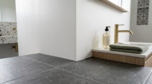 Close-up of textured charcoal grey porcelain floor tiles meeting a crisp white wall, featuring a low oak vanity with a brushed brass basin mixer tap, hand soap, folded waffle towel, and terrazzo wall accents in the background.