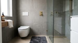 Modern wet room bathroom layout featuring a white wall-hung toilet with a dual flush plate beside a frameless glass shower enclosure fitted with brushed brass shower fixtures, large grey wall tiles, and wood-look flooring.