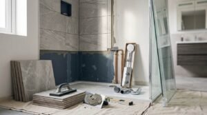 Shower installation in progress featuring a white acrylic shower base, partially tiled grey stone walls with a recessed niche, exposed blue waterproofing membrane, copper plumbing pipes, unboxed chrome shower fixtures, and tiling tools on a drop cloth.