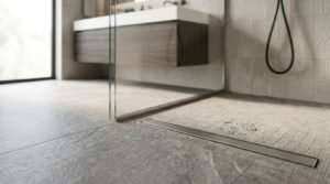 Close-up low-angle view of a curbless walk-in shower featuring slip-resistant textured grey stone floor tiles, water droplets near a brushed stainless steel linear drain, a frameless glass screen, and a floating dark wood vanity.