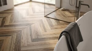 Chevron-patterned wood-look porcelain tile flooring in a modern bathroom, featuring water droplets inside a glass walk-in shower enclosure next to a freestanding bathtub with a grey towel.