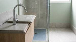 Bathroom renovation in progress showing a light oak vanity with stone countertop tile samples, a gooseneck chrome mixer tap, sage green walls, and a walk-in shower with waterproof membrane flooring.