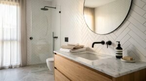 Modern bathroom renovation featuring a marble vanity, black wall-mounted faucet, round mirror, herringbone tile wall, and frameless glass shower