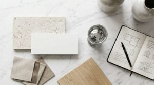 Renovation material selection board with limestone and subway tiles, fabric swatches, oak panel, polished chrome fixture, and architectural sketchbook on a marble table