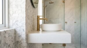 Luxury bathroom design featuring a white floating concrete benchtop, round vessel sink, brushed brass mixer tap, Carrara marble mosaic wall tiles, and glass shower enclosure