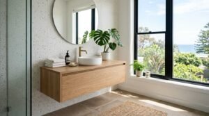 Modern floating oak bathroom vanity with round vessel sink, brushed brass faucet, terrazzo feature wall, and coastal window view