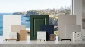 Kitchen material mood board set against a Sydney coastal cliff and ocean backdrop, showcasing white, forest green, deep navy, and beige cabinet profiles, rattan cane webbing, marble, terrazzo, and stone samples, paired with antique brass, brushed brass, and matte black handles.