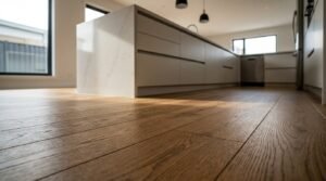 Low-angle perspective of wide-plank engineered oak timber flooring in a sunlit modern kitchen, extending towards a handleless minimalist island with a white marble quartz waterfall benchtop and pendant lighting.