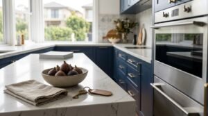 Modern renovated kitchen ready for property sale featuring a Calacatta quartz waterfall island styled with fresh figs in a ceramic bowl, house keys on a leather fob, linen napkin, deep navy shaker cabinets with brass hardware, and built-in stainless steel double ovens.