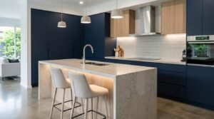 Modern open-plan kitchen featuring a grey-veined quartz waterfall island with under-bench LED strip lighting and two upholstered bar stools, paired with matte navy blue flat-panel cabinetry, light timber upper cupboards, white subway tile splashback, and three white pendant lights.