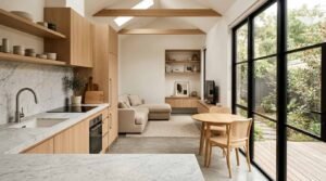 Interior view of a modern open-concept granny flat featuring vaulted ceilings with exposed wood beams, a marble kitchen countertop with light oak cabinets, and black-framed glass sliding doors opening to a garden patio