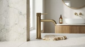 Close-up of a deck-mounted brushed brass basin mixer tap with knurled handle detail beside a travertine stone slab sample on a white terrazzo countertop