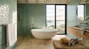 Luxury coastal bathroom featuring glossy seafoam-green subway tiles, a freestanding stone bathtub, frameless glass shower, floating wood vanity with brass fixtures, and floor-to-ceiling windows showcasing an ocean view and natural coastal landscape.