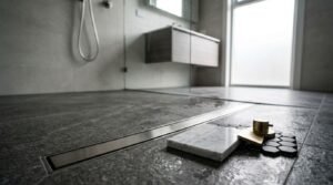 Modern wet room with a linear floor drain, dark stone tiles, glass shower screen, and material samples in the foreground.