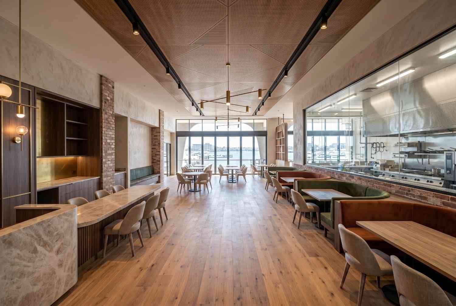 Modern restaurant interior fit-out featuring custom booth seating, open commercial kitchen, timber flooring, and waterfront views. Contemporary hospitality design showcasing premium dining spaces, stylish finishes, functional restaurant layout, and inviting customer experience for upscale food and beverage venues