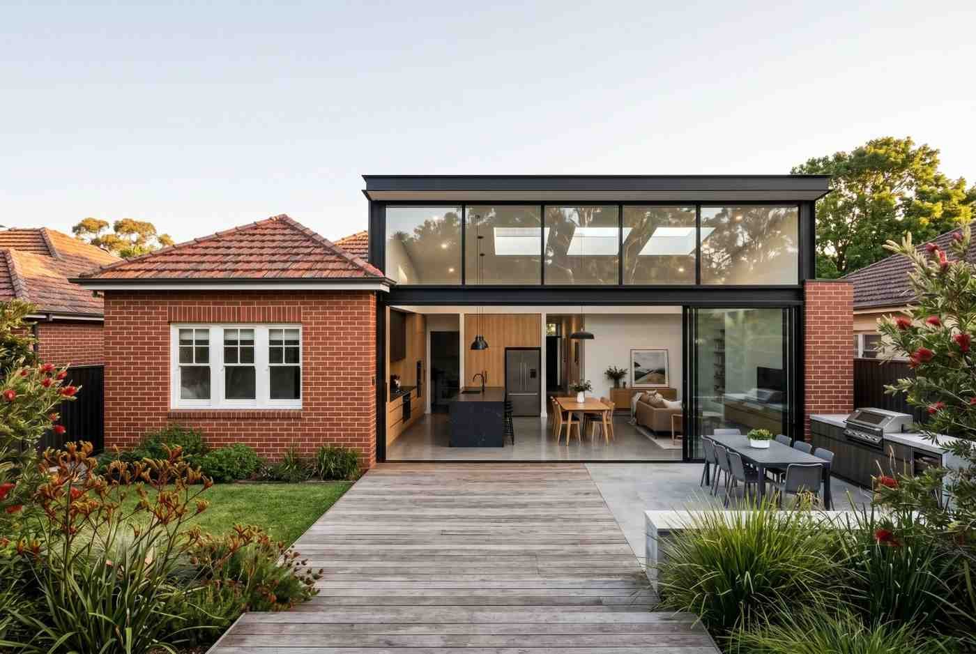 Modern home extension featuring floor-to-ceiling glass, open-plan kitchen and dining area, landscaped backyard, and seamless indoor-outdoor living. Contemporary residential renovation combines traditional brick architecture with modern design, maximizing natural light, functionality, energy efficiency, and outdoor entertainment space.