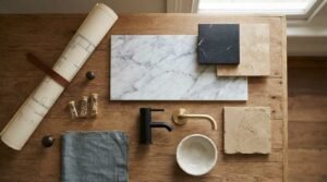 Flat lay bathroom material palette flat lay featuring rolled architectural blueprints, Carrara marble slab, black marble tile, travertine swatches, matte black tap, and brushed brass spout on a wooden table