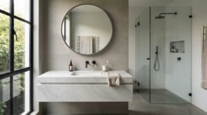 Minimalist ensuite bathroom featuring a floating white marble vanity, wall-mounted matte black tapware, circular wall mirror, and a glass-enclosed walk-in shower next to a black-framed window