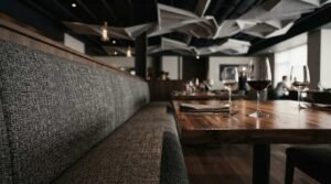 Modern restaurant interior with acoustic ceiling panels, wooden dining tables, upholstered booth seating, and wine glasses in a contemporary dining space