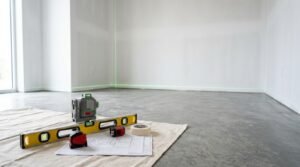 Laser level, spirit level, tape measure, painter's tape, and floor plan arranged on a drop cloth inside an empty room prepared for renovation.