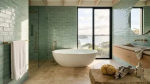 Luxury coastal bathroom featuring glossy seafoam-green subway tiles, a freestanding stone bathtub, frameless glass shower, floating wood vanity with brass fixtures, and floor-to-ceiling windows showcasing an ocean view and natural coastal landscape