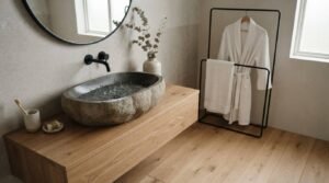 Modern Japandi bathroom renovation with a natural stone vessel sink, floating oak vanity, matte black wall-mounted faucet, round mirror, light wood flooring, and a minimalist robe and towel rack.