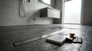Modern wet room with a linear floor drain, dark stone tiles, glass shower screen, and material samples in the foreground