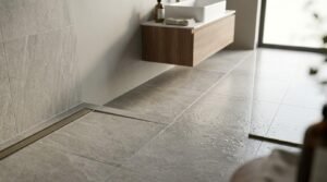 Open-plan wet room with curbless shower floor, brass linear trench drain, textured grey stone-look porcelain tiles, and a floating wood vanity