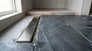 Stainless steel linear drain channel set in thin-set mortar next to large dark slate floor tiles with tile spacers during shower installation