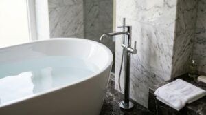 White oval freestanding bathtub filled with clear water next to a floor-mounted chrome mixer faucet in a marble-tiled bathroom