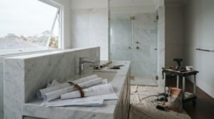 Luxury bathroom renovation under construction featuring architectural blueprints, chrome basin mixer tap, and marble vanity island with frameless glass shower enclosure and tiling tools