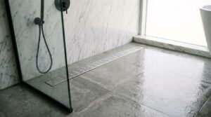 Stainless steel slotted linear drain set flush in wet grey porcelain floor tiles inside a walk-in shower with marble walls