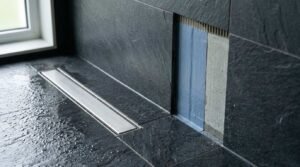 Stainless steel linear shower drain in dark slate tiles with a cutaway wall section showing blue waterproofing membrane and mortar layer