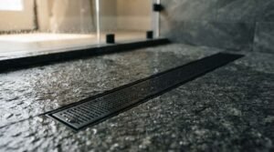 Matte black linear shower drain recessed into a wet textured dark granite stone shower floor with water droplets