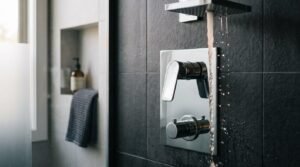 Wall-mounted polished chrome thermostatic shower mixer control panel with running water flowing against dark slate wall tiles