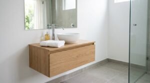 Wall-hung light oak single-drawer floating vanity with a round white ceramic vessel basin, tall chrome mixer tap, and frameless rectangular mirror beside a glass walk-in shower