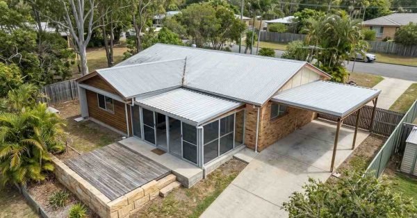 Aerial view of a residential property compliance inspection showing the home exterior, roof condition, carport, driveway, deck, boundary fencing, landscaping, and overall site layout for assessment and documentation.