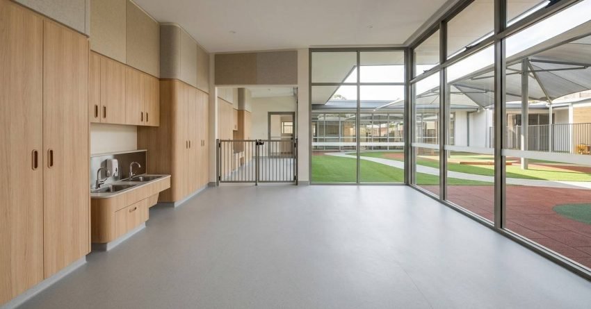 Modern childcare centre renovation interior featuring custom timber cabinetry, integrated child-height sink station, expansive floor-to-ceiling glazing, secure gated transition area, durable commercial flooring, acoustic wall panels, abundant natural daylight, and direct connection to a landscaped outdoor play courtyard in a newly completed early learning facility