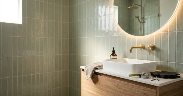 Modern bathroom vanity with a white vessel sink, brushed brass wall-mounted tapware, round backlit LED mirror, and timber cabinetry against sage green vertical subway tile walls.