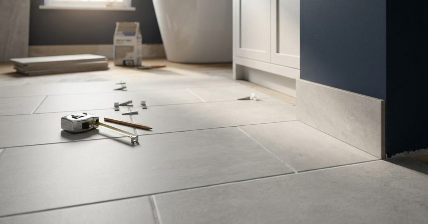 Primary Alt Text Bathroom floor renovation in progress featuring newly laid large-format light grey tiles with leveling clips, a measuring tape, and a pencil in the foreground, leading toward a white freestanding bathtub and vanity against a dark navy wall.