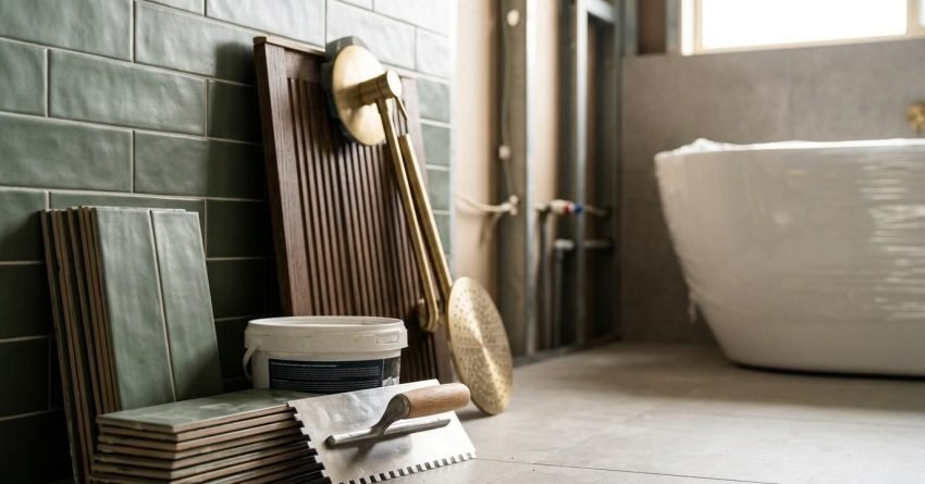 Bathroom renovation materials including sage green subway tiles, a notched trowel, tile adhesive tub, fluted timber vanity panel, and brushed brass rainfall showerhead next to an uninstalled freestanding bathtub.