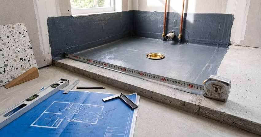 Waterproofed shower base pan with liquid membrane coating, brass drain, measuring tape, and architectural blueprints on concrete floor