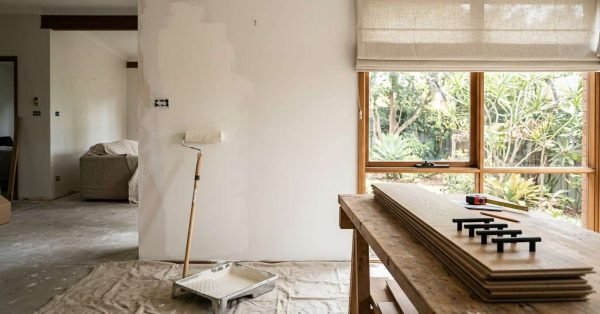 Living room under renovation featuring a paint roller resting against a partially painted white wall, paint tray on a canvas drop cloth, and timber flooring planks with black hardware on a workbench