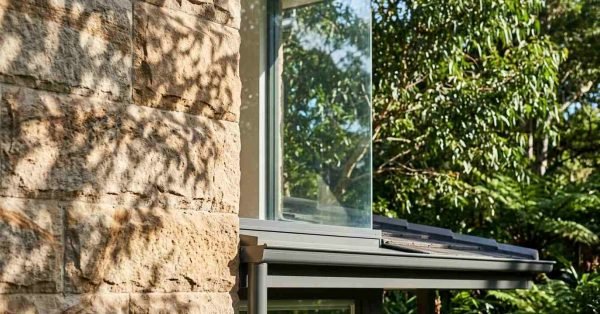 Detailed architectural exterior featuring rough-hewn sandstone block cladding, frameless glass corner window, and charcoal metal awning guttering framed by green foliage
