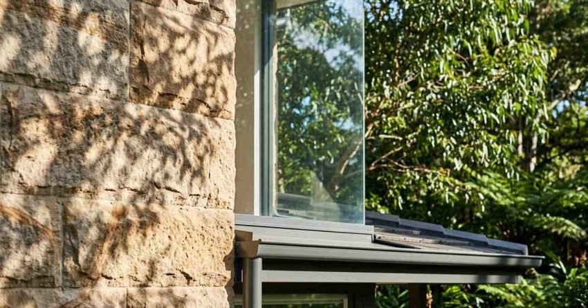 Detailed architectural exterior featuring rough-hewn sandstone block cladding, frameless glass corner window, and charcoal metal awning guttering framed by green foliage