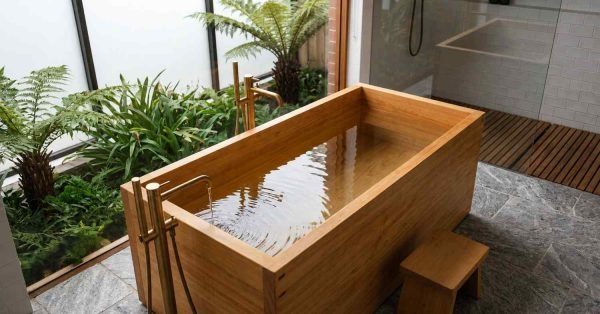 Rectangular wooden Japanese soaking tub filled with water featuring floor-mounted brushed brass faucets beside an indoor garden courtyard